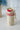 Mug Térmico Germ x Coca-Cola Vintage 700ml / 23 Oz Mug térmico Coca-Cola vintage color crema con tapa y acento rojo, asa de tela clara, en un ambiente interior con flores azules al fondo.